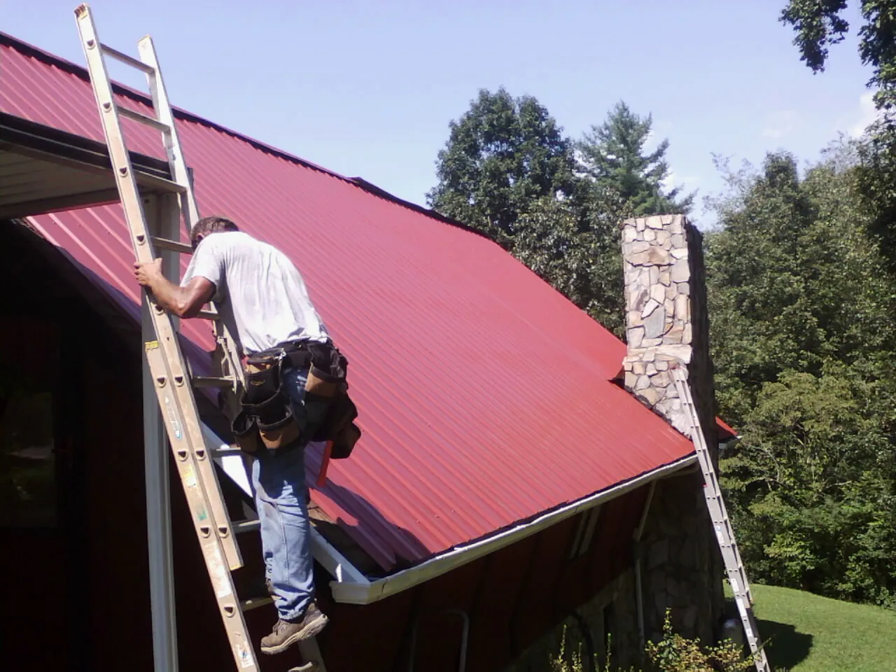 Roofer on a ladder beside a red metal roof and stone chimney — Soffit & Fascia Repair by Roofing Brothers in Hillburn